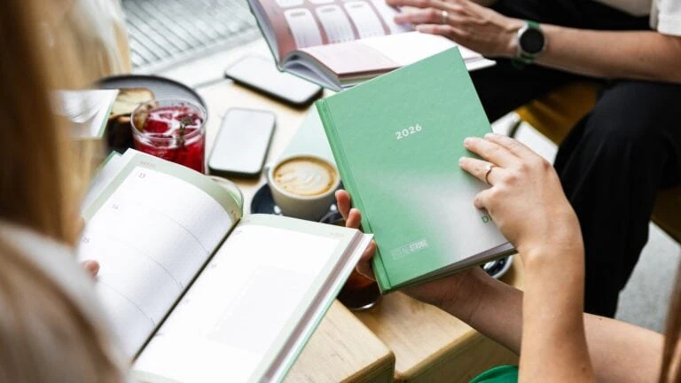 Osoby držící plánovače s rokem 2026, káva a občerstvení na stole, příprava na nový rok.