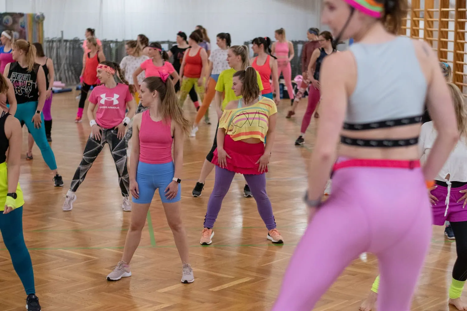 Skupina žen v barevných sportovních oblečeních se účastní energického tréninku v tělocvičně.