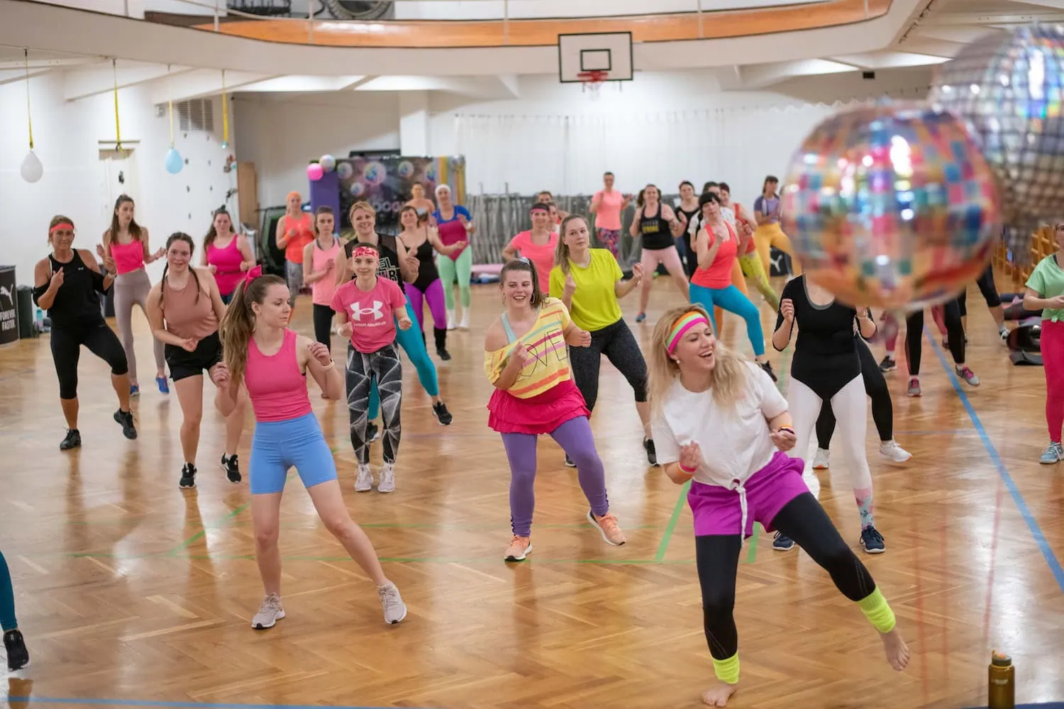 Skupina žen v barevném sportovním oblečení tancuje na lekci aerobiku v tělocvičně.