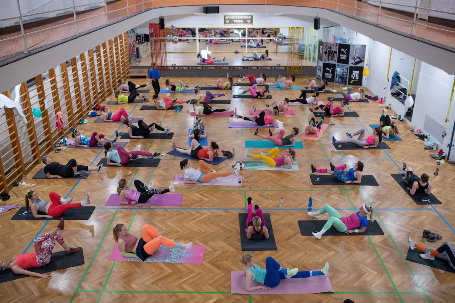 Skupina lidí cvičí na podložkách v tělocvičně, většina v barevných sportovních oblečeních.