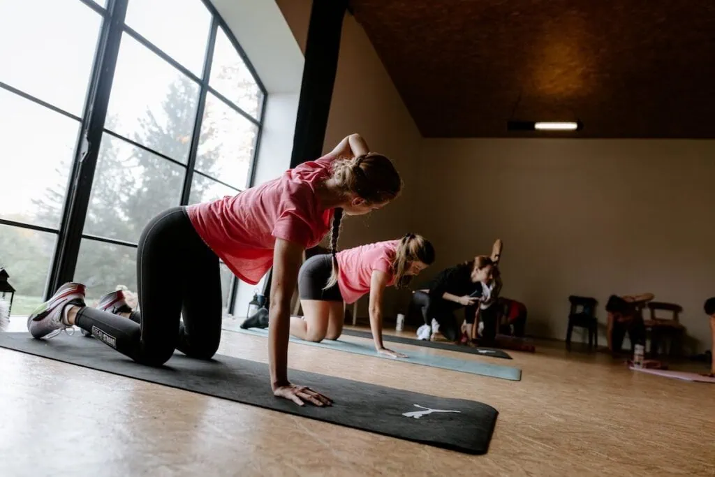 Ženy v růžových tričkách cvičí na podložkách v tréninkové lekci v prostorné místnosti.