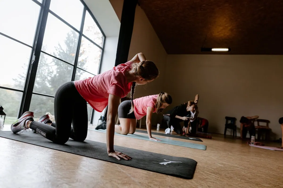 Ženy v růžových tričkách cvičí na podložkách v tréninkové lekci v prostorné místnosti.