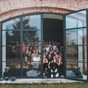 Skupina žen se usmívá a pózuje u velkého okna, v pozadí přírodní prostředí a tréninkový prostor.