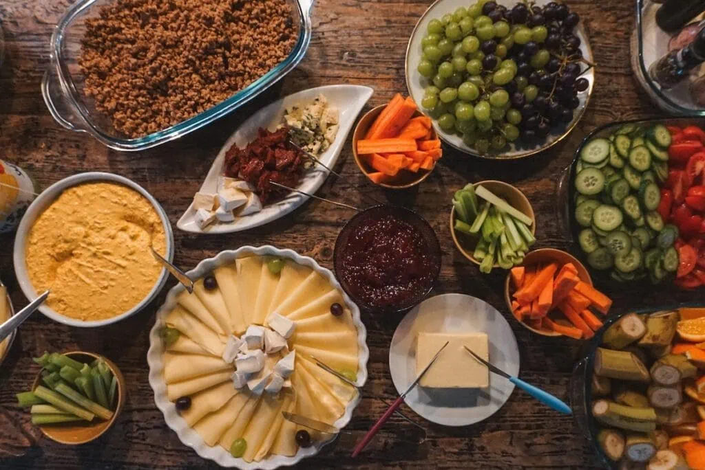Stůl plný různých pokrmů: zelenina, ovoce, pomazánky a sýry, připravené na servírování.