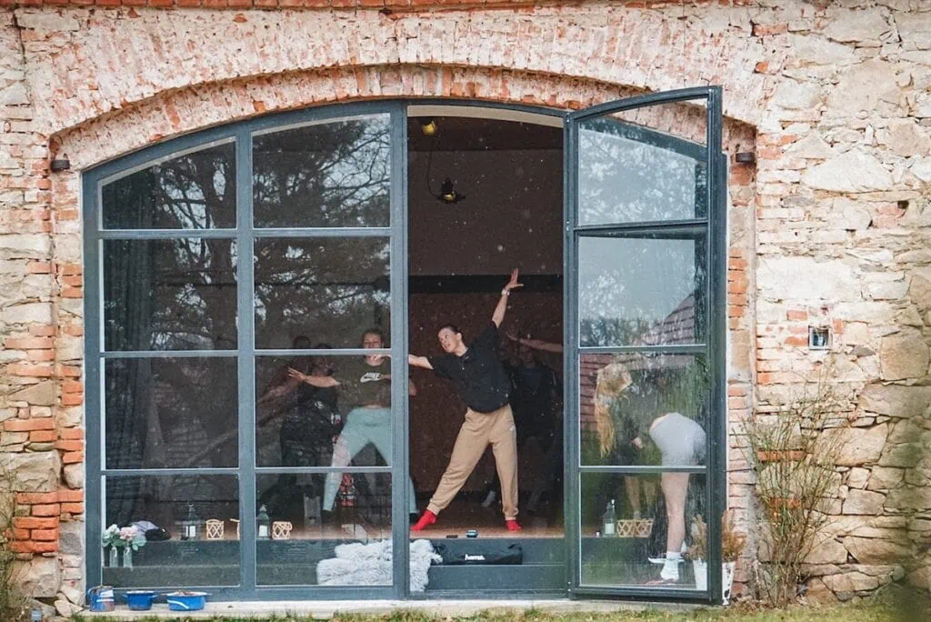 Skupina lidí cvičí v místnosti s velkými okny, vidět také květiny a cvičební vybavení.