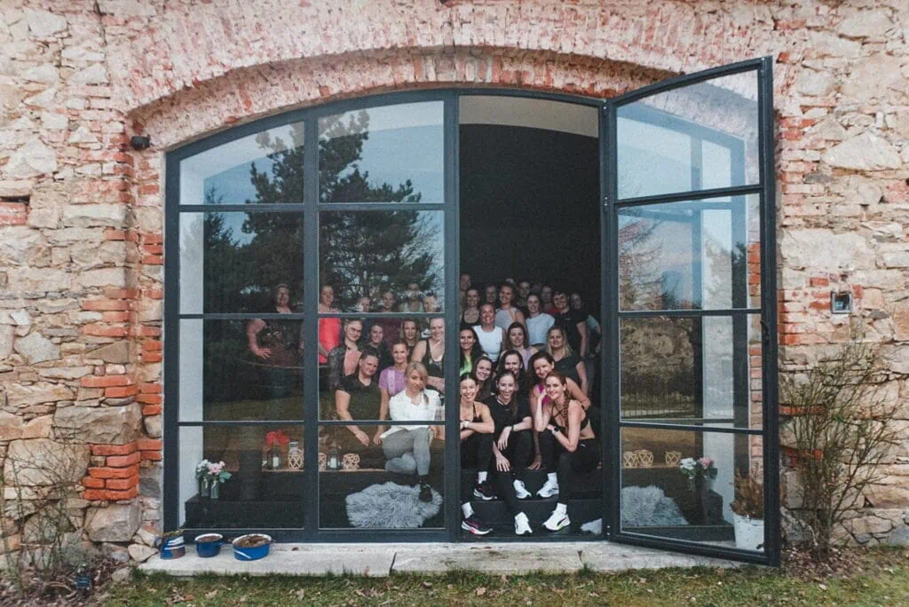 Skupina žen se usmívá a pózuje před otevřenými dveřmi historické budovy během fitness víkendu.