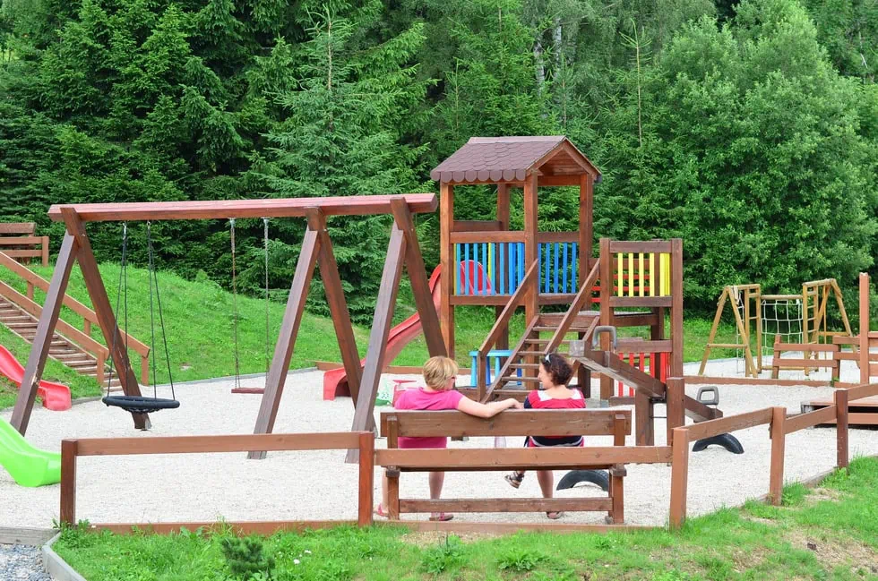 Dvě děti sedí na lavičce vedle dětského hřiště s houpačkami a skluzavkou v zeleném prostředí.