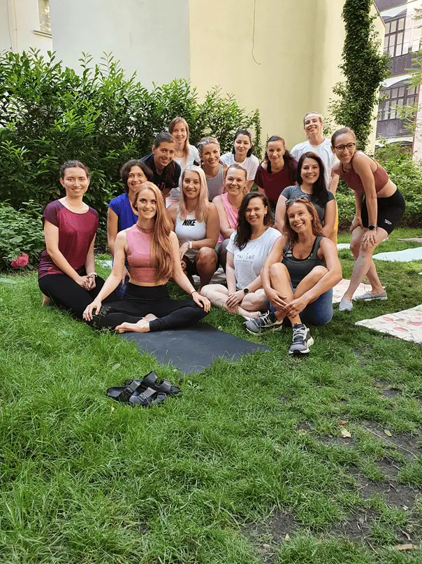 Skupina lidí na tréninkové lekci v přírodě, sedící na podložkách na trávě, usmívají se.