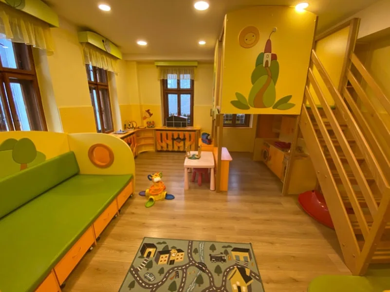 Hravý interiér dětského koutku s barevnými stěnami, zelenou sedačkou, hračkami a dřevěným schodištěm.