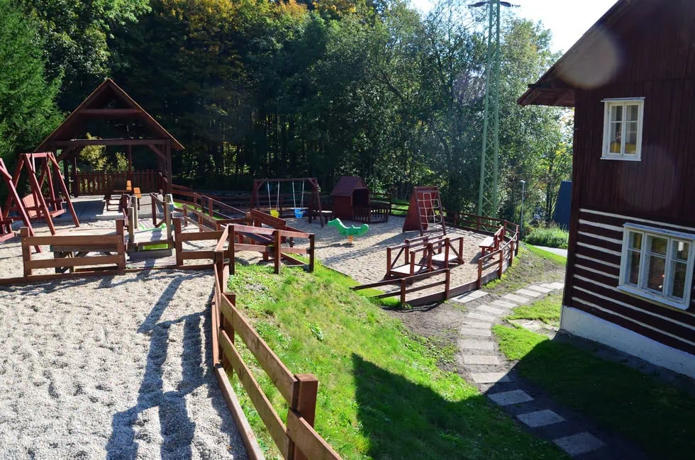 Hřiště s dřevěnými prolézačkami, houpačkami a pískovištěm obklopené zelení a dřevěným plotem.