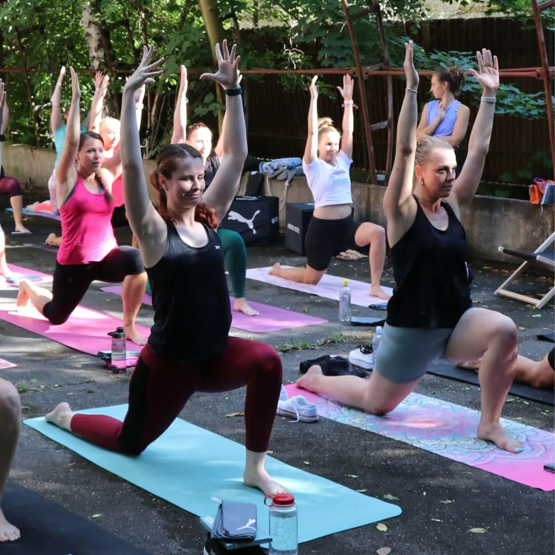 Skupina lidí cvičí na podložkách, některé mají ruce vztyčené, v pozadí stromy a cvičební pomůcky.