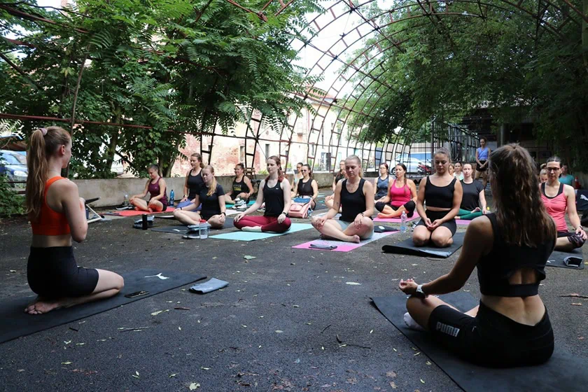 Skupina žen cvičí jógu pod vzrostlými stromy v otevřeném prostoru.