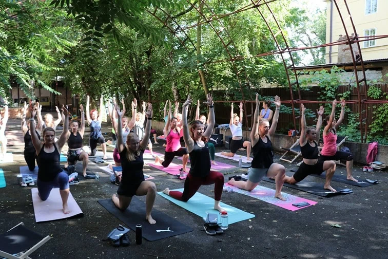 Skupina lidí cvičících na podložkách v parku, provádějících různé pozice s rukama vztyčenýma nad hlavou.