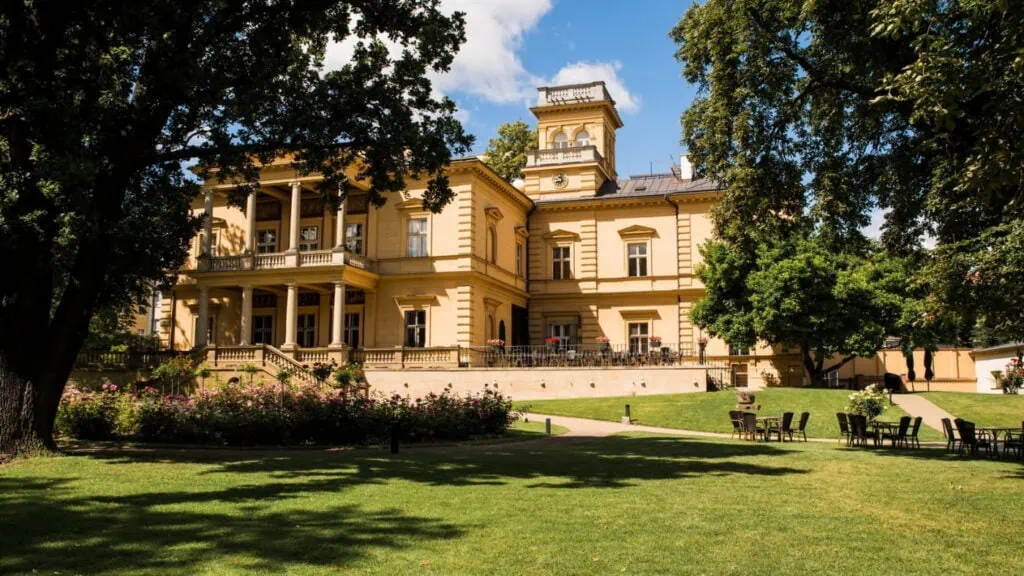 Elegantní budova s terasou obklopená zelení a květinami, ideální pro venkovní lekce a tréninkové aktivity.